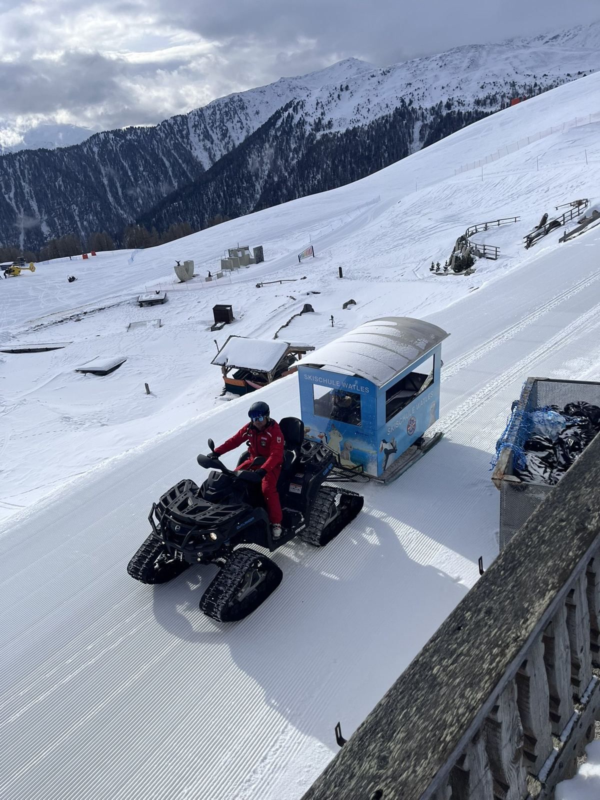 Skilehrer im roten Anzug fährt mit einem Raupen-Quad den blauen Kinderwagen der Skischule Watles über die Piste