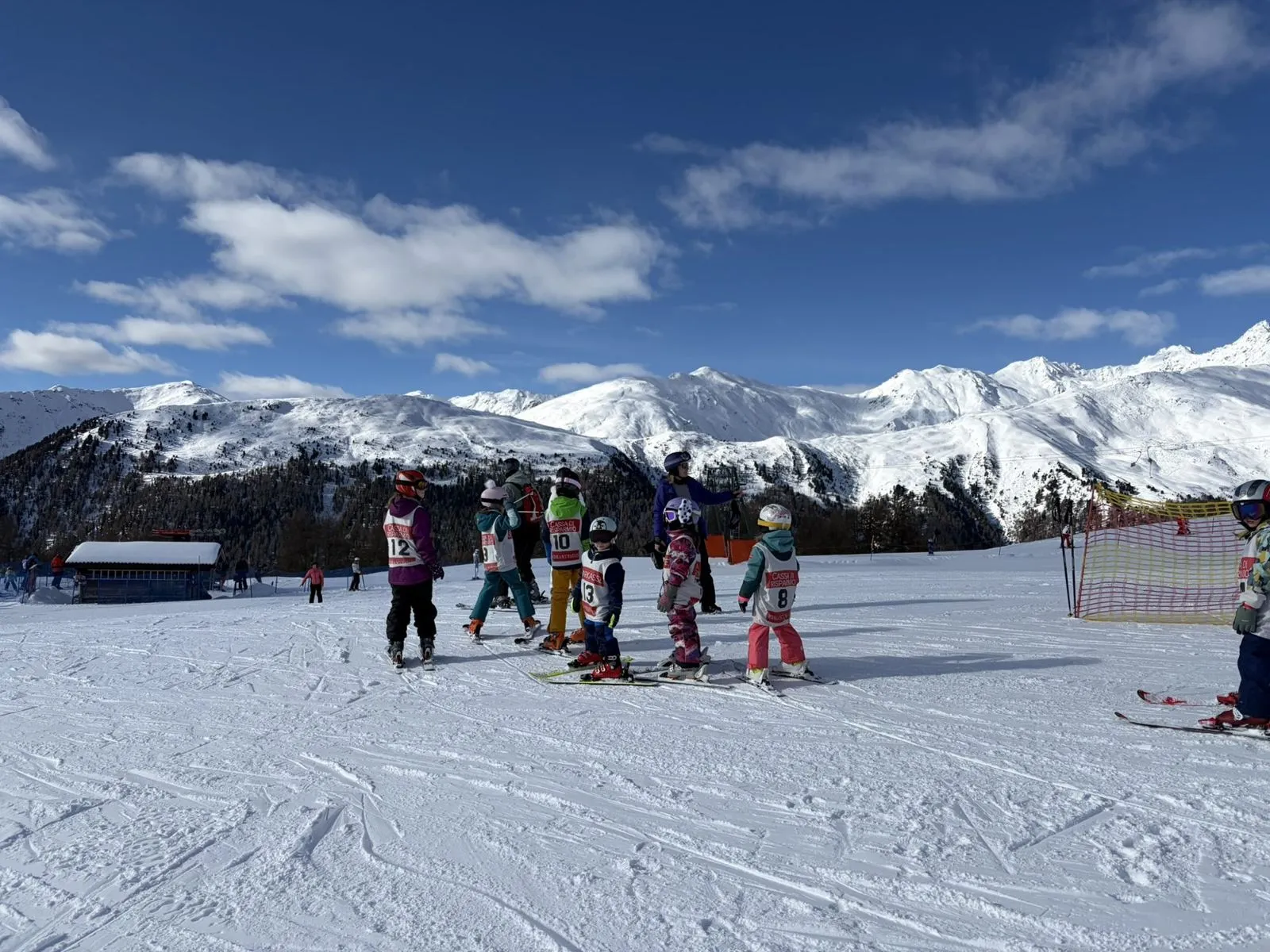 Kindergruppe mit Startnummern und Skilehrerin auf der Piste, im Hintergrund verschneite Berge